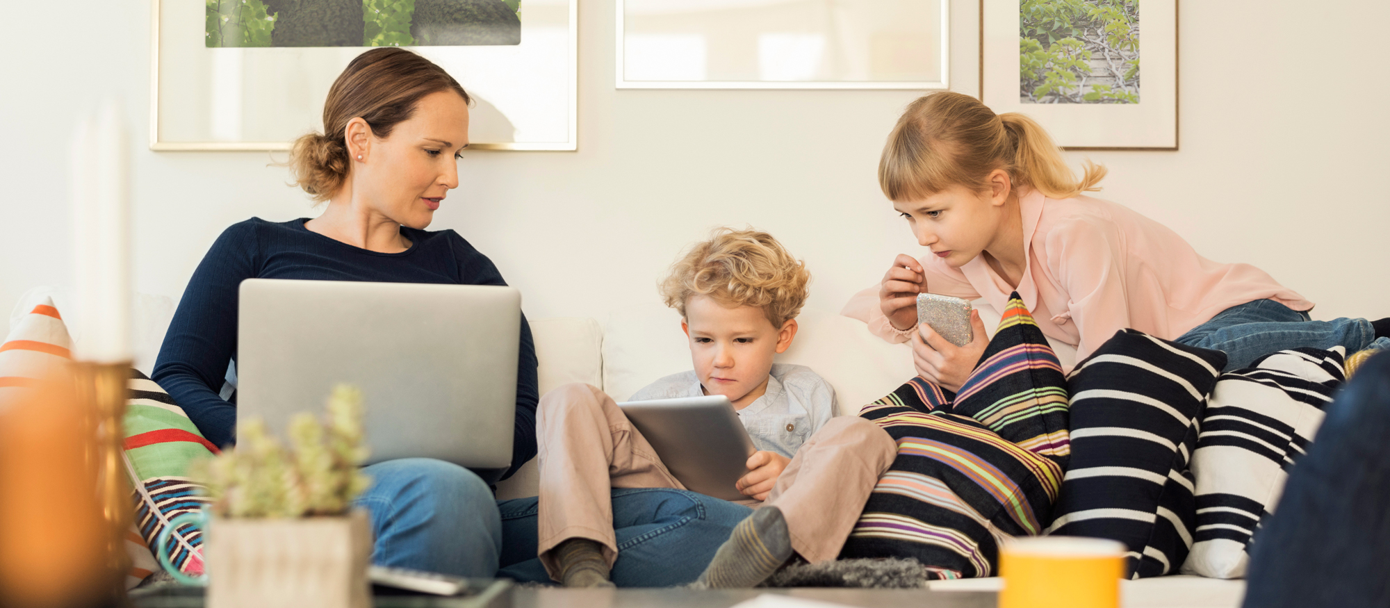 Mamma och två barn sitter i soffa och tittar på olika skärmar